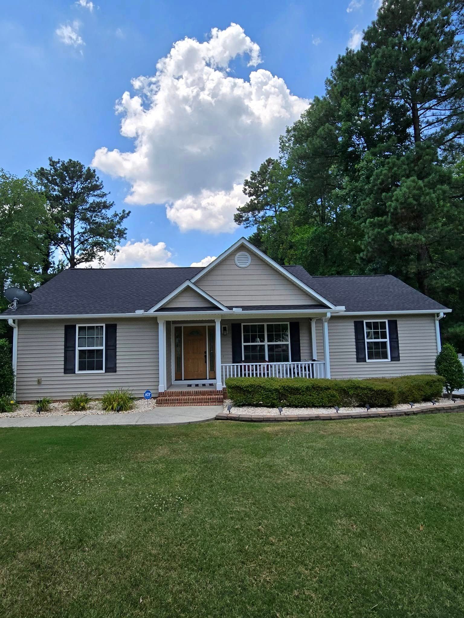 A single-story gray house with a peaked roof, a front porch, dark shutters, and a lawn under a blue, cloudy sky.