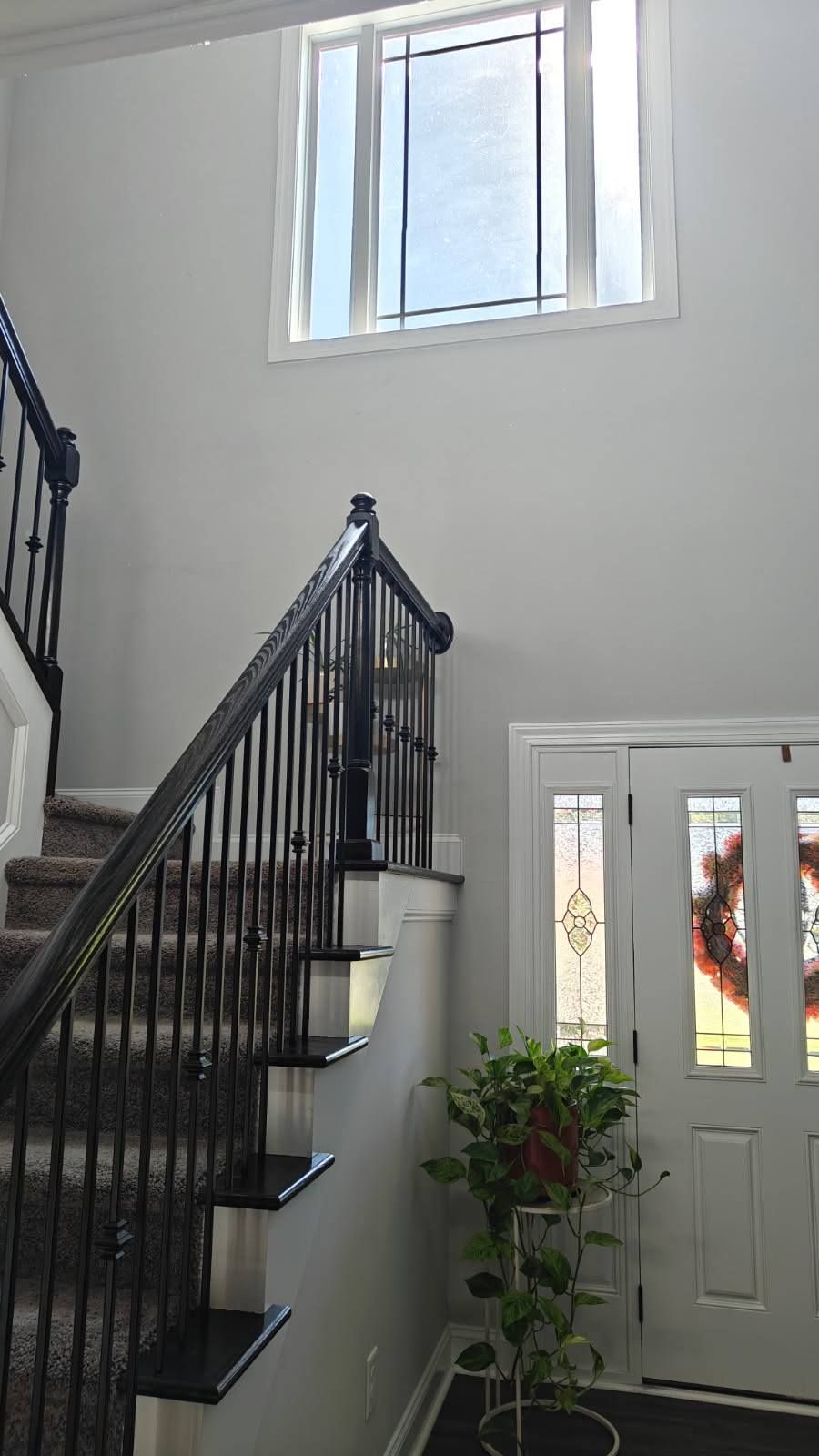 An interior foyer featuring a staircase with black railings, gray walls, a tall window, and a white front door.