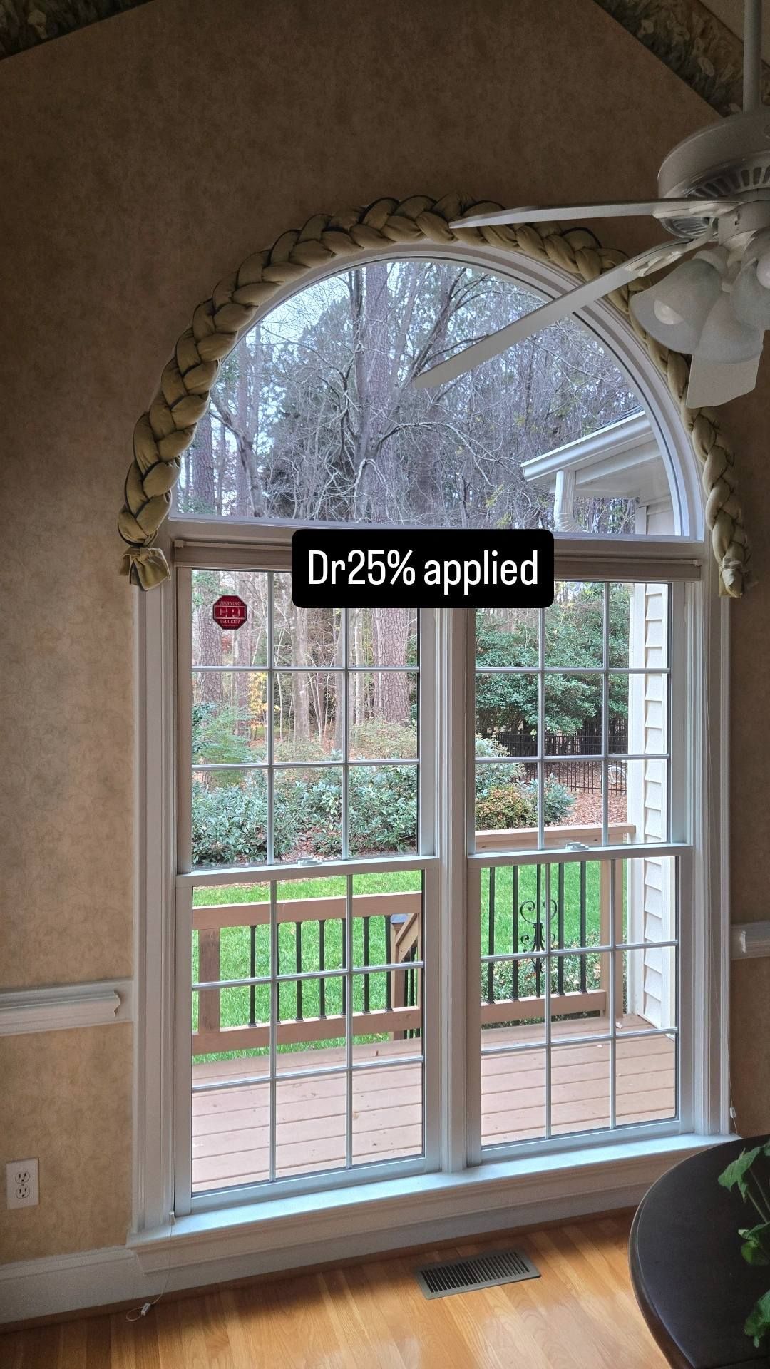 Large arched window in a room with tan wallpaper and a ceiling fan, looking out onto a wooden porch and trees.
