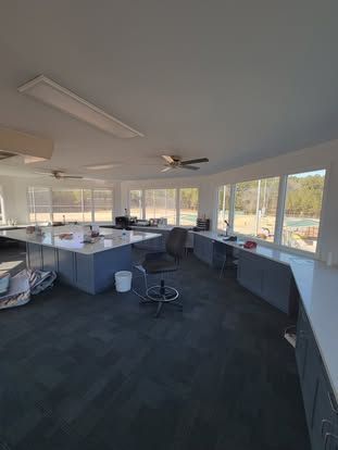 A bright office space with large windows, dark grey carpet, white desks, and a black office chair in the center.