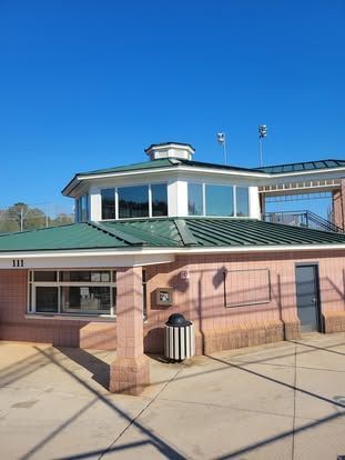 A pink brick building with a green metal roof, a cupola, and a trash can on a concrete plaza under a clear blue sky.