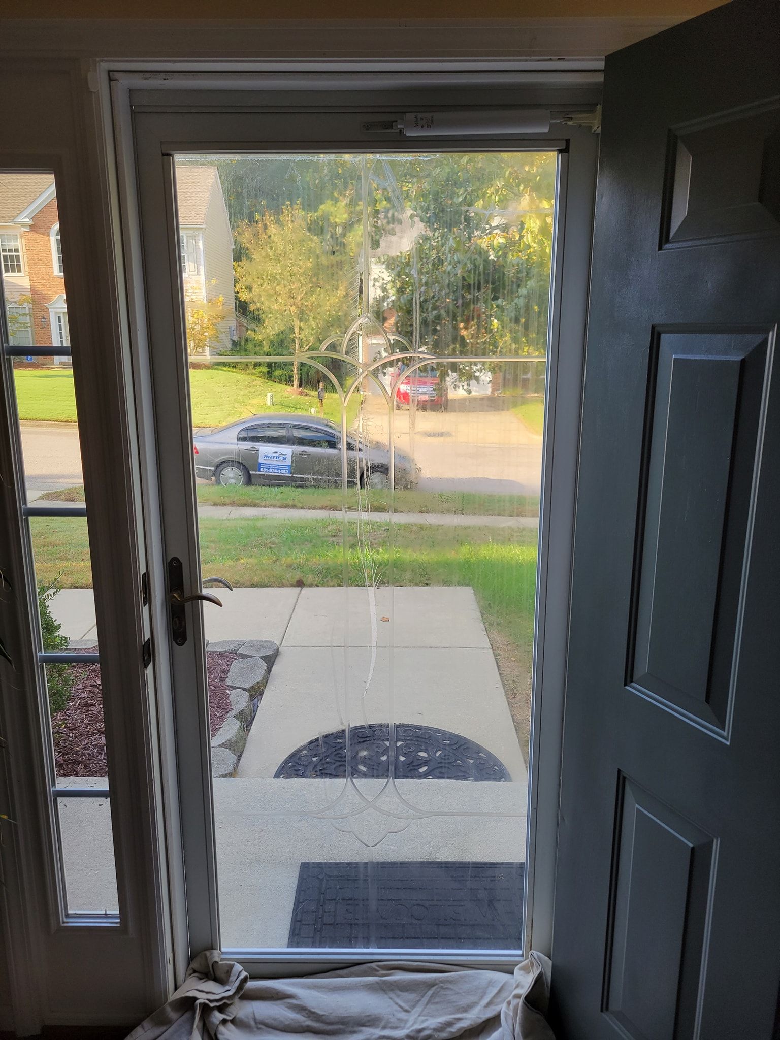 A storm door with a large, vertical crack in the glass pane, viewed from inside a house with a dark main door open.