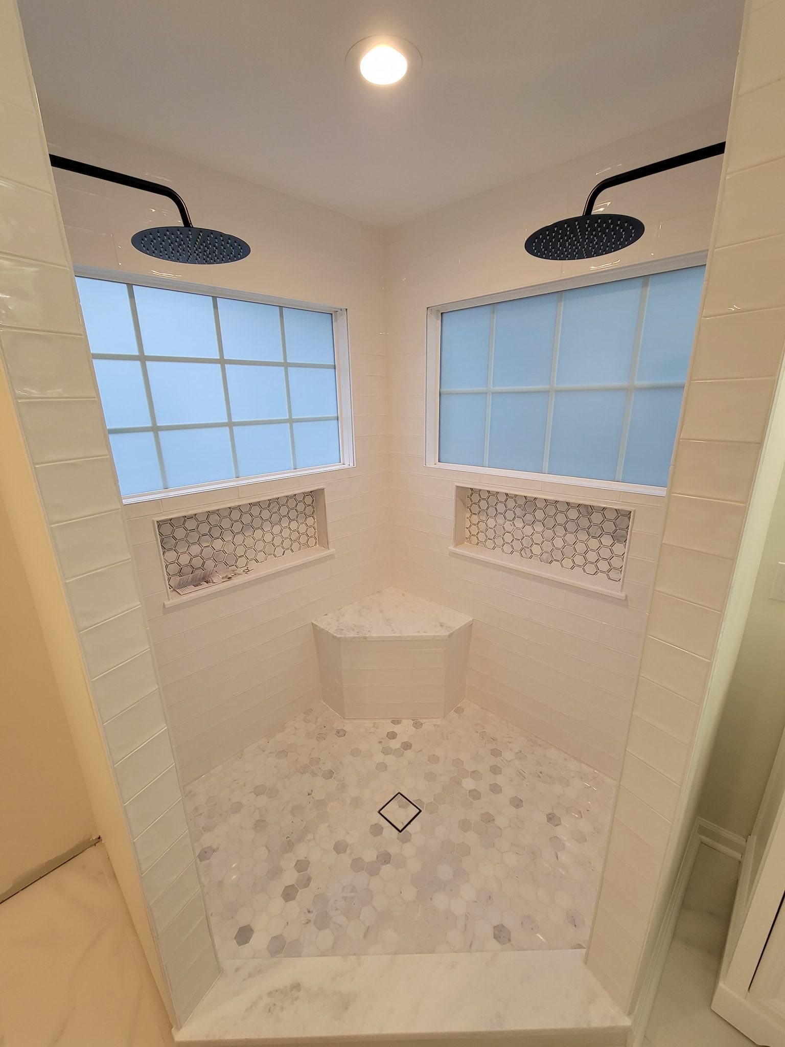 A modern corner shower with dual black showerheads, white tile walls, mosaic niches, a marble seat, and patterned tile floor.