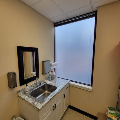 A stainless steel sink with a granite countertop, a mirror, and a large, frosted-glass window in a small, neutral-toned room.
