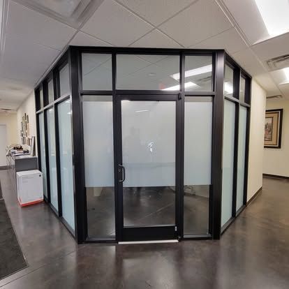 A modern office glass enclosure with black metal frames and privacy-frosted windows, featuring a central glass door.