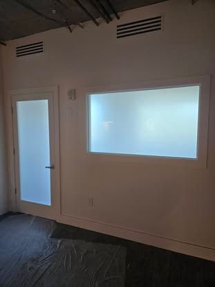 An interior office wall featuring a white door and a large window, both with frosted glass, below exposed ceiling pipes.