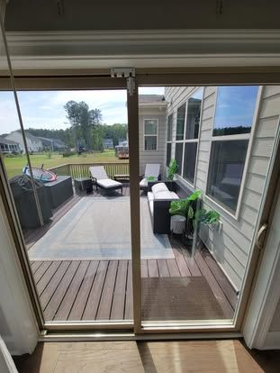 View of a wooden outdoor deck with lounge chairs and a sofa on a rug, seen through a sliding glass door.