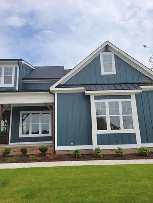 A modern blue-sided house with white trim, a metal roof, and a small flower bed in front of a green lawn under a blue sky.