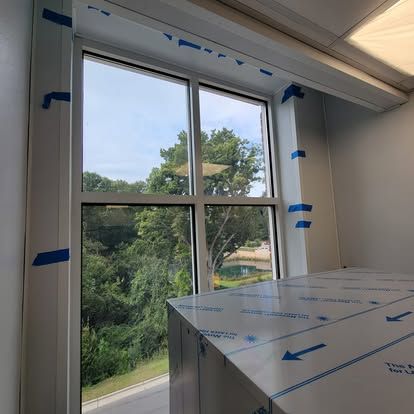 A window view of trees and a pond, with blue painter's tape on the white window frame and construction materials inside.
