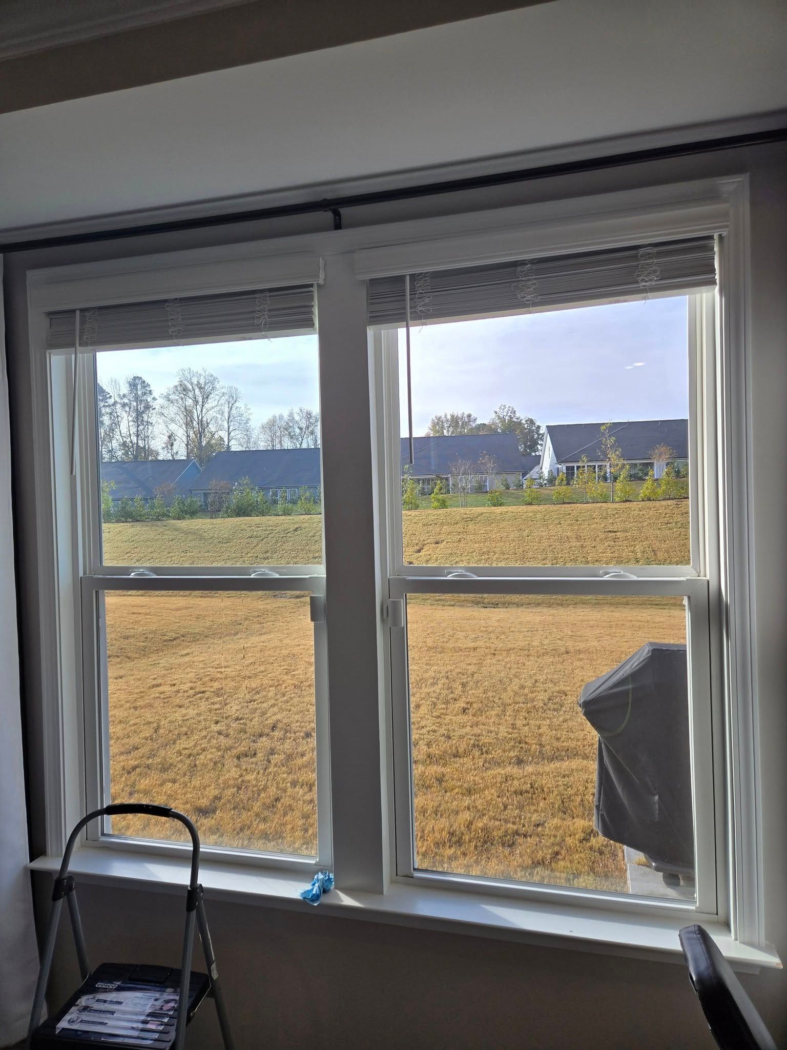 Two windows in a room with white trim, gray blinds, and a view of a grassy field and houses under a bright sky.