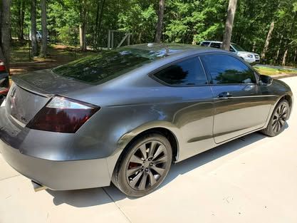 A gray two-door Honda Civic coupe parked on a concrete driveway with a wooded background.