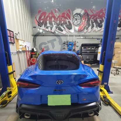 A blue Toyota Supra parked in a garage between two yellow and blue hydraulic vehicle lifts.