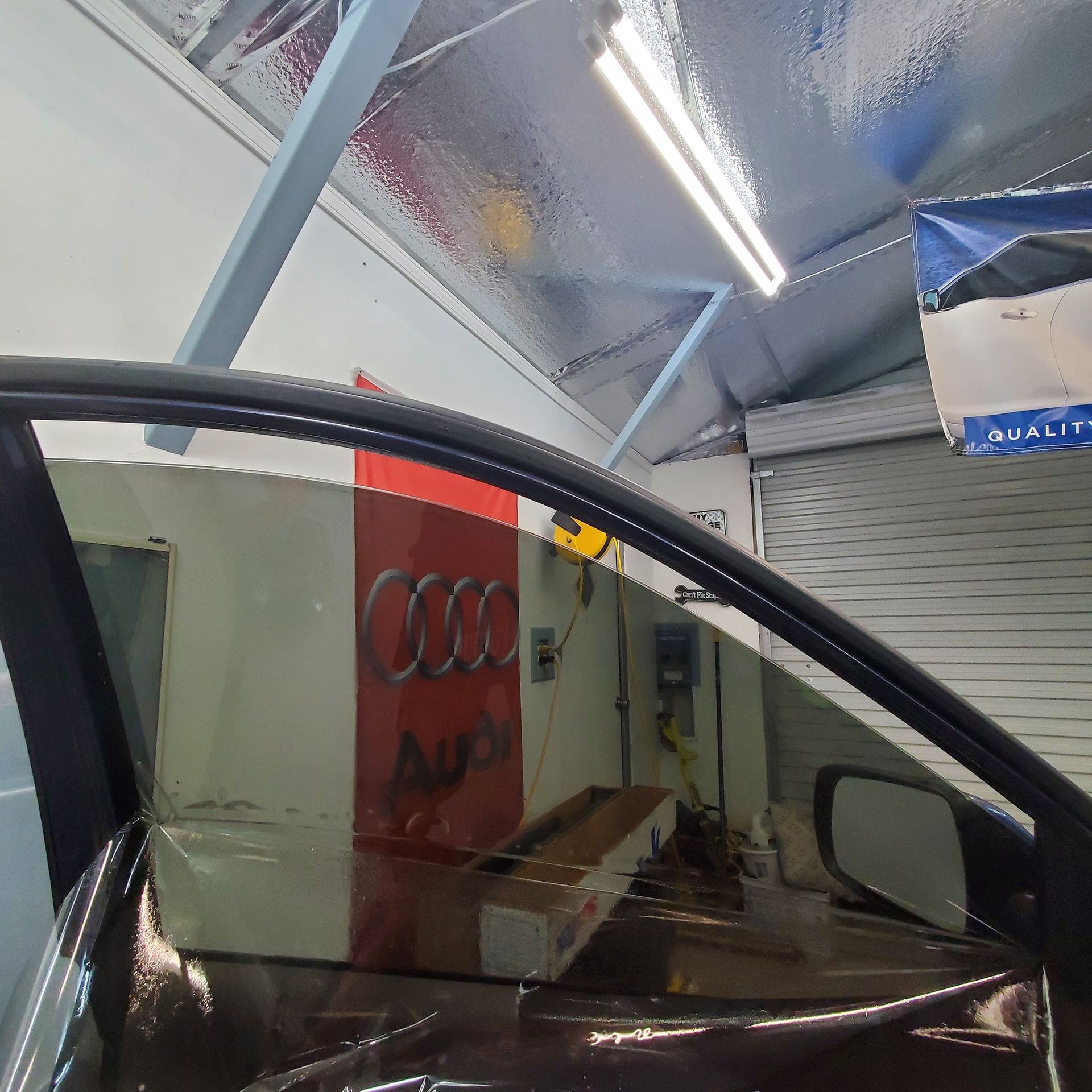 A view from inside an Audi, showing a tinted car window being installed in an automotive shop setting.