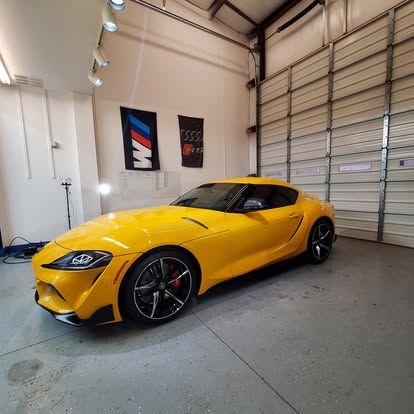A bright yellow Toyota Supra parked inside a clean garage with white walls and a large, metal roll-up door.
