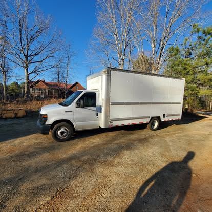 A white box truck parked on a gravel lot under a clear blue sky, with trees and a house in the background.