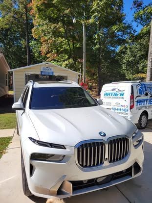 A white BMW SUV parked in a driveway next to a white work van with business branding, with a small building in the back.
