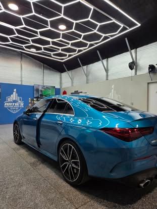 A teal Mercedes-Benz CLA parked in a bright studio with a hexagonal LED ceiling light pattern.