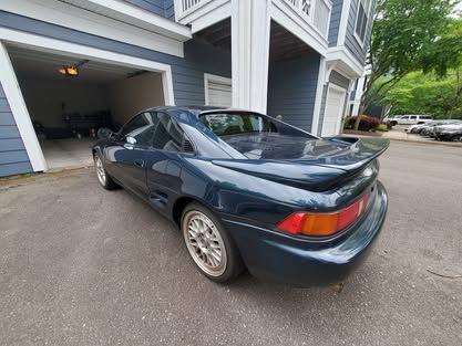 A teal Toyota MR2 parked in a driveway in front of a house.