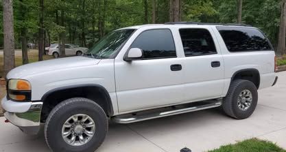 A white GMC Yukon XL SUV parked on a concrete driveway in a wooded, outdoor setting.