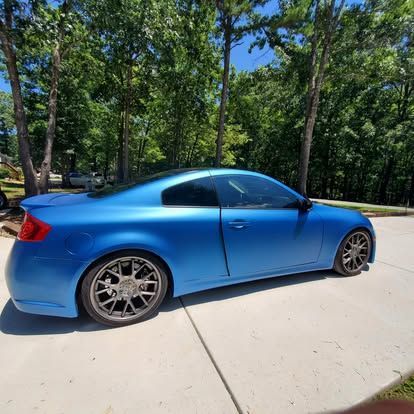 A matte blue Infiniti G35 coupe parked on a concrete driveway surrounded by trees on a sunny day.