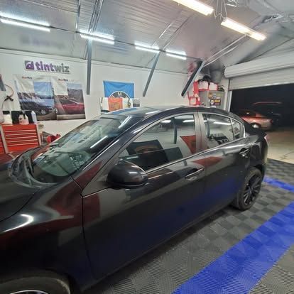 A dark sedan parked in a garage with a tiled floor, TintWiz sign, and car-themed decor on the walls.