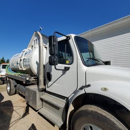 A white industrial vacuum tanker truck parked next to a white building under a clear blue sky.