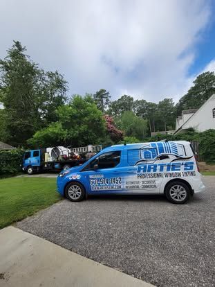 A blue and white work van and a utility truck parked on a gravel driveway in front of a house and trees.