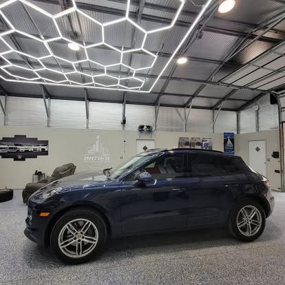 A dark blue Porsche SUV parked in a garage with a hexagonal LED ceiling light grid and speckled grey floor.