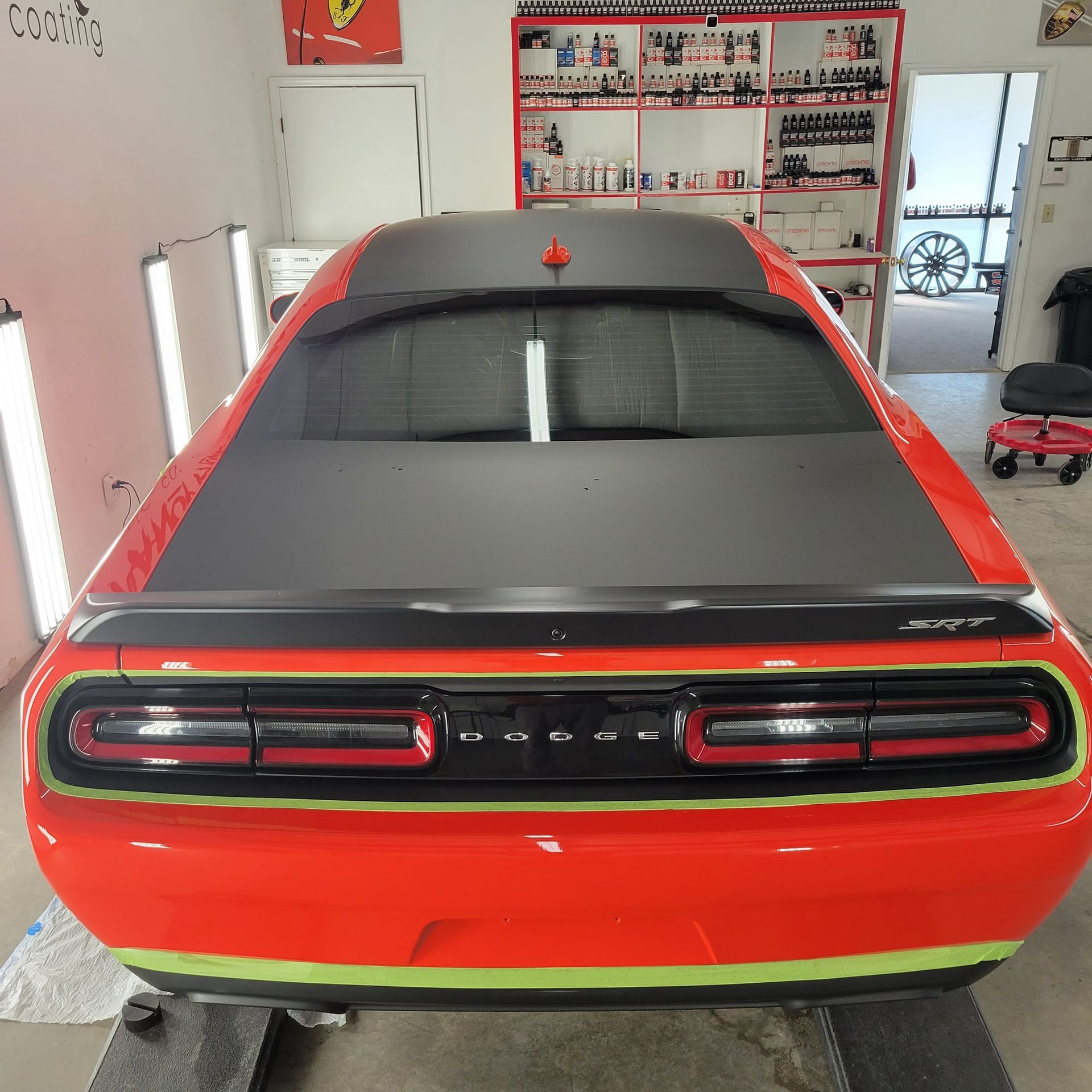 Rear view of a bright orange Dodge Challenger parked in an auto garage, featuring black accents and masking tape.
