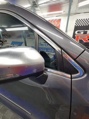 A side view of a gray car's exterior, focusing on the silver side mirror and the chrome window trim in a garage setting.