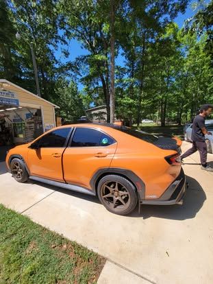 An orange Subaru WRX with bronze wheels and black accents is parked on a concrete driveway near a building and trees.