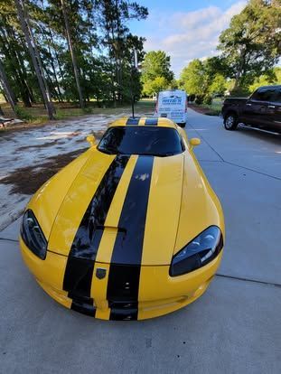 A yellow Dodge Viper with black racing stripes parked on a paved driveway near trees and a truck.