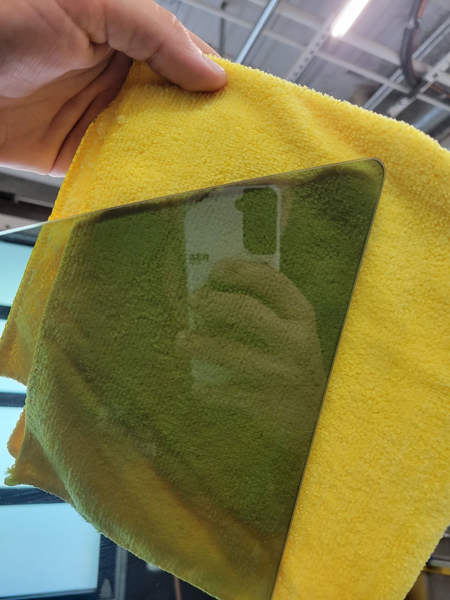 A hand holds a yellow microfiber cloth over a reflective, dark-tinted window, revealing a reflection of a person filming.