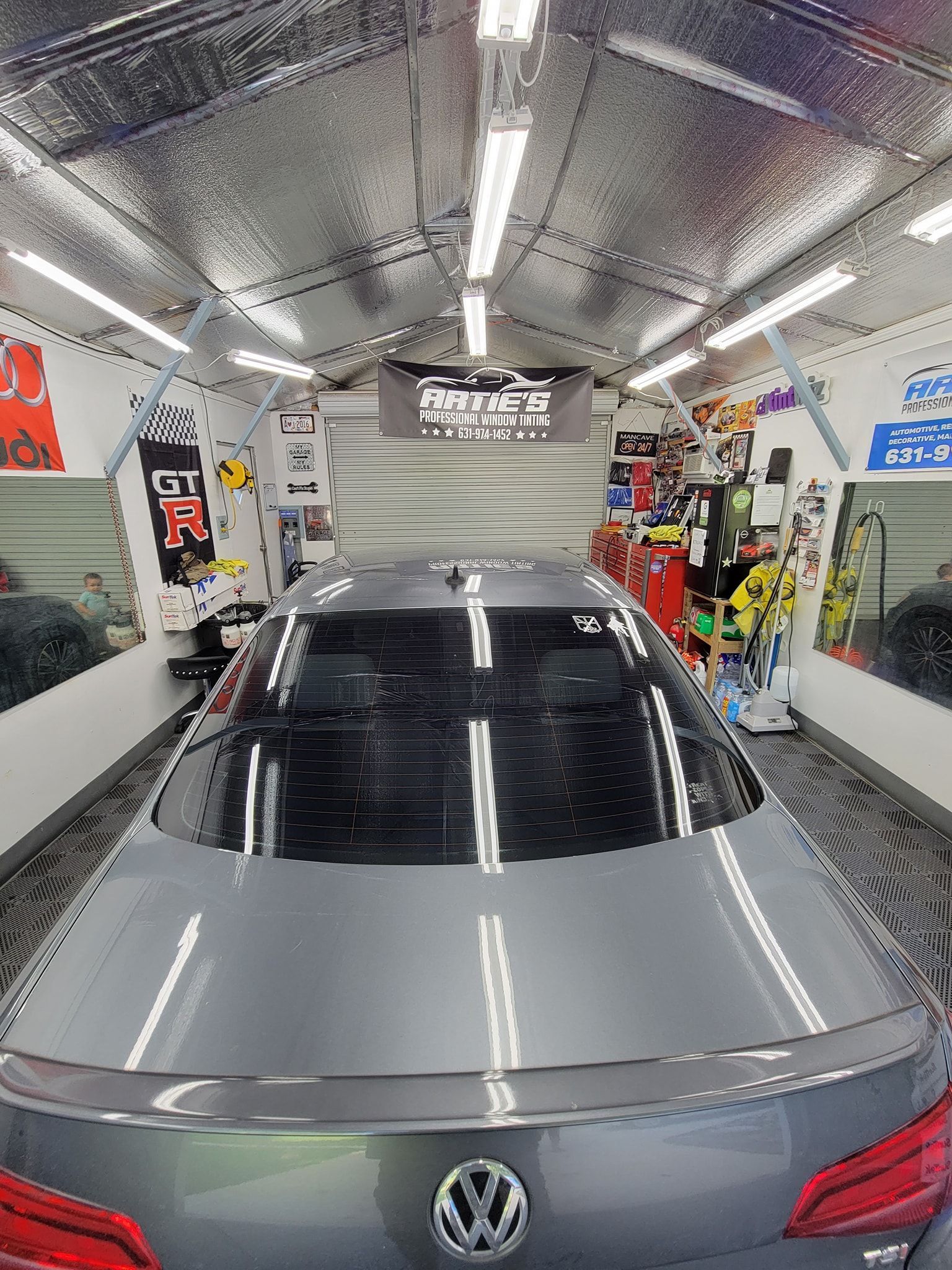 A gray Volkswagen parked in a narrow, well-lit garage with metallic reflective walls and various wall decals.