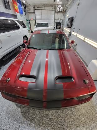 A close-up of a deep red car with wide grey racing stripes on the hood, parked inside a brightly lit garage.