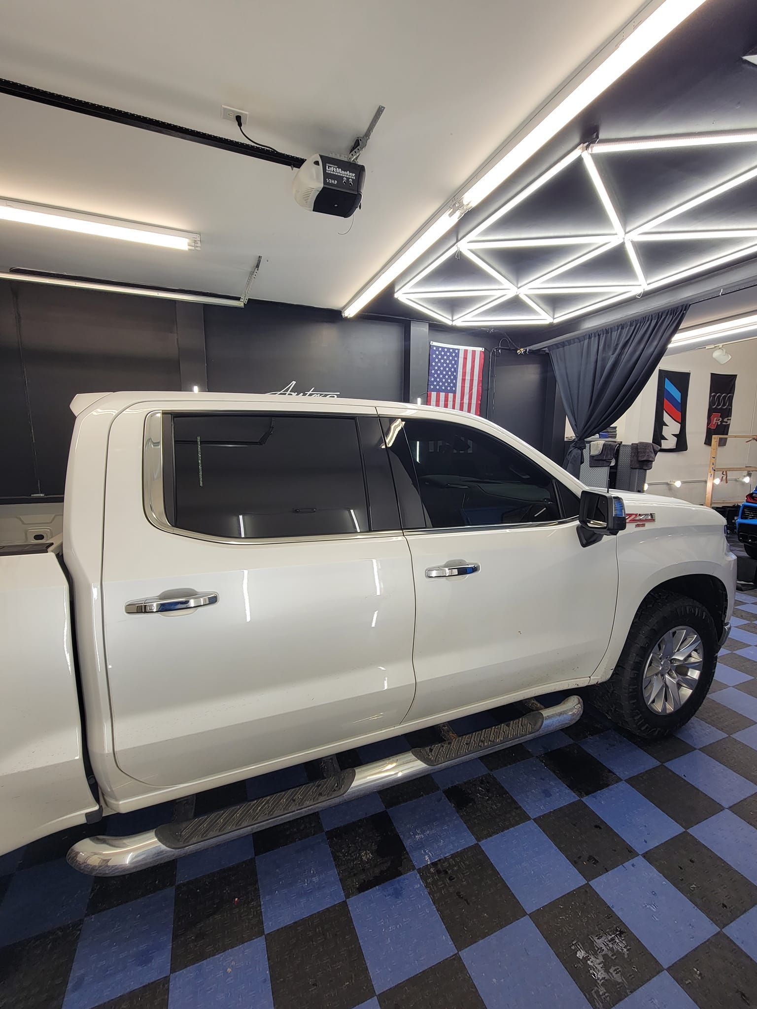 A white crew cab pickup truck parked inside a garage with patterned floor tiles and bright overhead lighting.