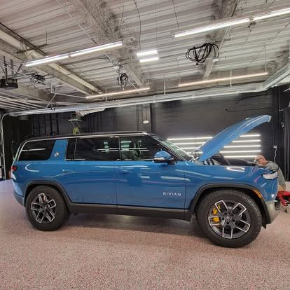 A blue Rivian SUV with its hood open in a garage, where a person is working under the hood.