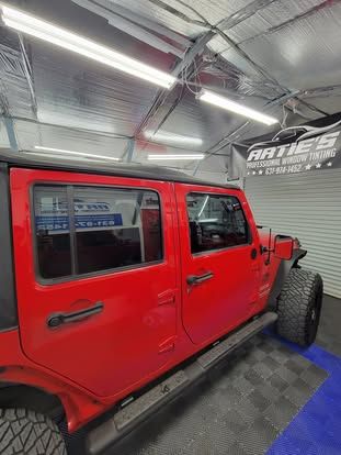 A side profile of a bright red Jeep Wrangler parked inside a brightly lit automotive garage with a workshop sign overhead.