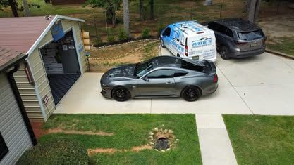 A gray sports car is parked in a residential driveway next to a shed and two other vehicles on a sunny day.