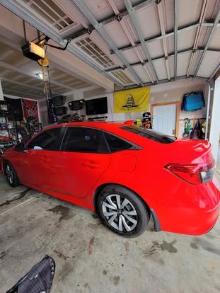 A bright red sedan parked inside a cluttered residential garage with a yellow Gadsden flag hanging on the back wall.