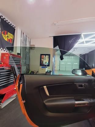 Open door of an orange car in a garage with a Porsche flag on the wall and a side-view mirror visible through the window.