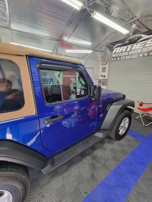 A blue Jeep Wrangler with a tan soft top parked inside a garage with grey flooring and overhead lighting.