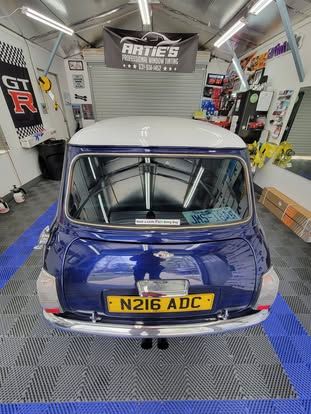 A blue vintage Mini Cooper with a white roof is parked inside a garage, viewed from the rear.