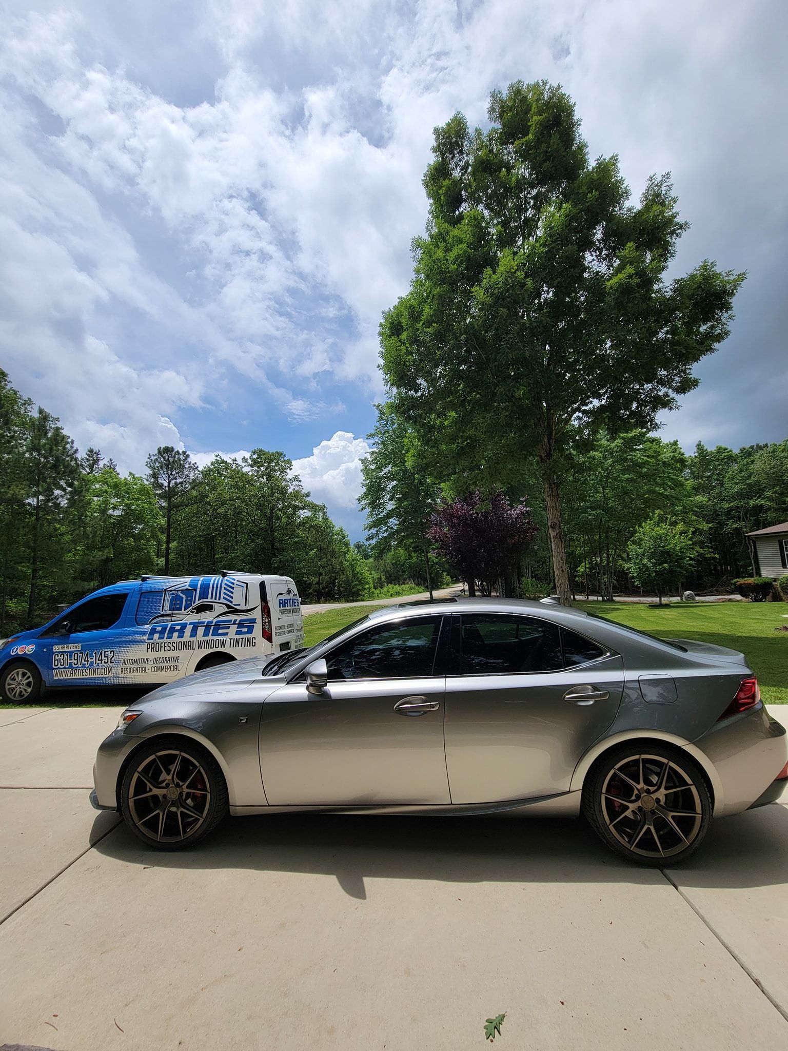 A metallic silver sedan parked on a concrete driveway next to a blue service van, with trees and a cloudy sky behind.