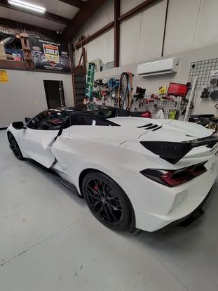 A white Chevrolet Corvette convertible with black wheels and a rear spoiler parked inside an automotive workshop.