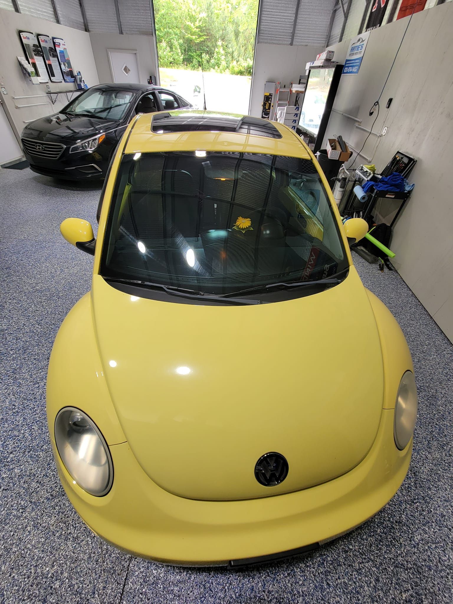 A yellow Volkswagen Beetle parked indoors on a speckled gray floor, with another car visible in the background.
