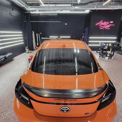 Rear view of an orange Toyota GR86 parked in a brightly lit professional detailing shop with black walls.