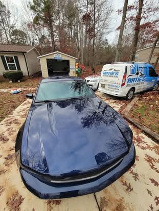 A dark blue sports car parked on a paved driveway in front of a house and a white work van with graphics.