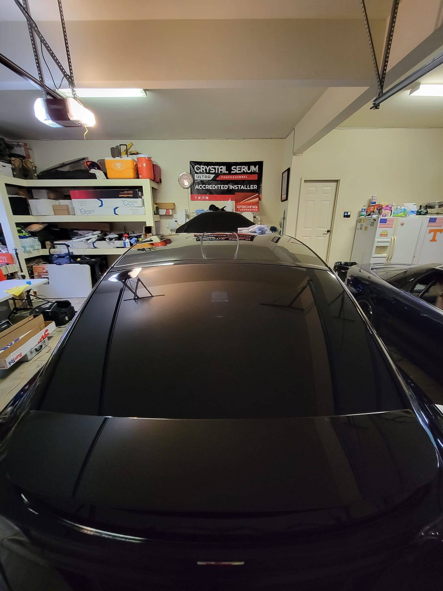 A high-angle view of a black car parked inside a cluttered garage with overhead lighting and storage shelves.