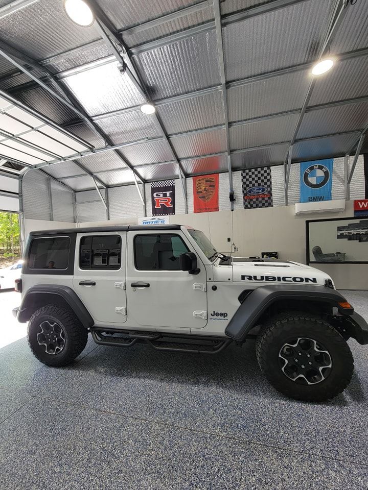 A white Jeep Wrangler Rubicon parked inside a well-lit garage with a speckled floor and automotive banners on the wall.